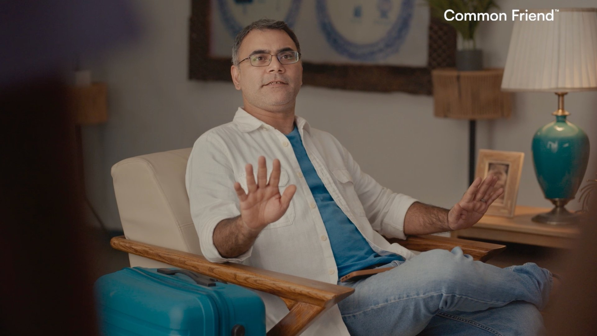 Male character seated beside blue suitcase in beach farmhouse interior designed for commercial ad film.