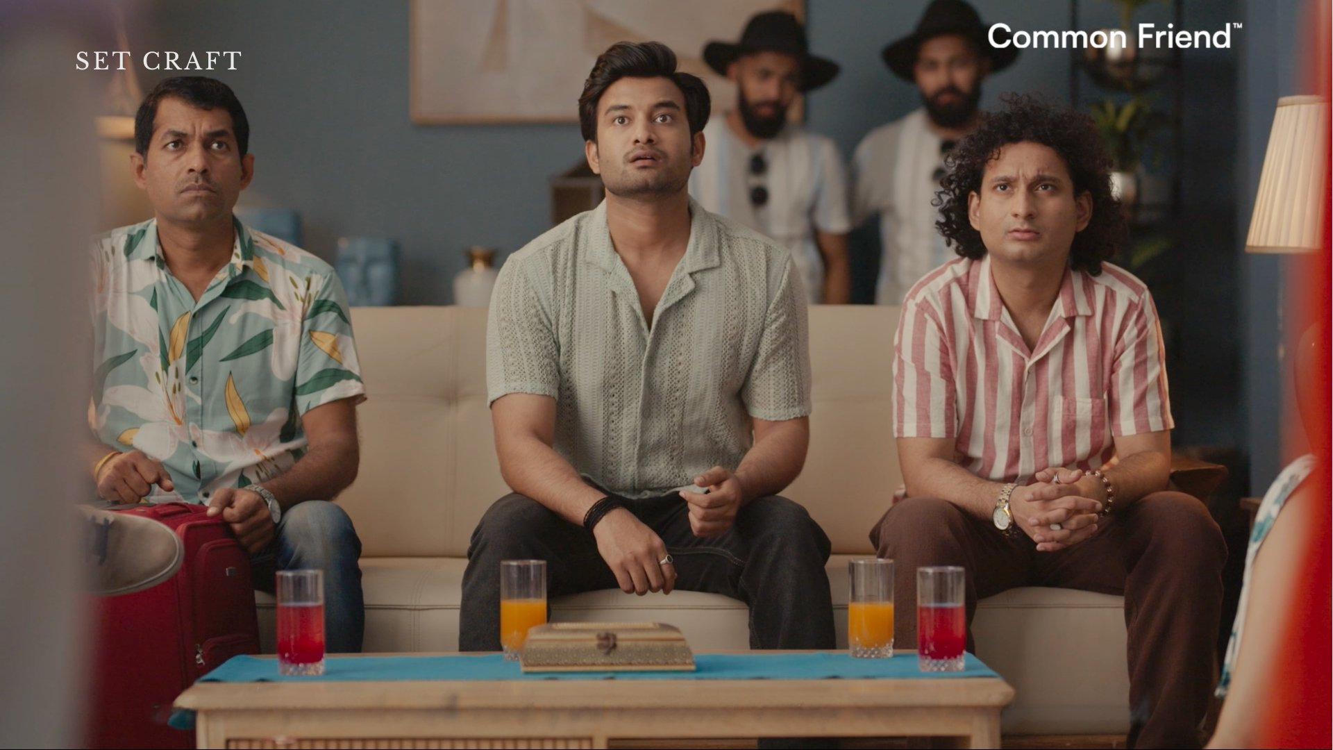 Three male characters seated on sofa in beach farmhouse commercial set with teal wall and layered props.