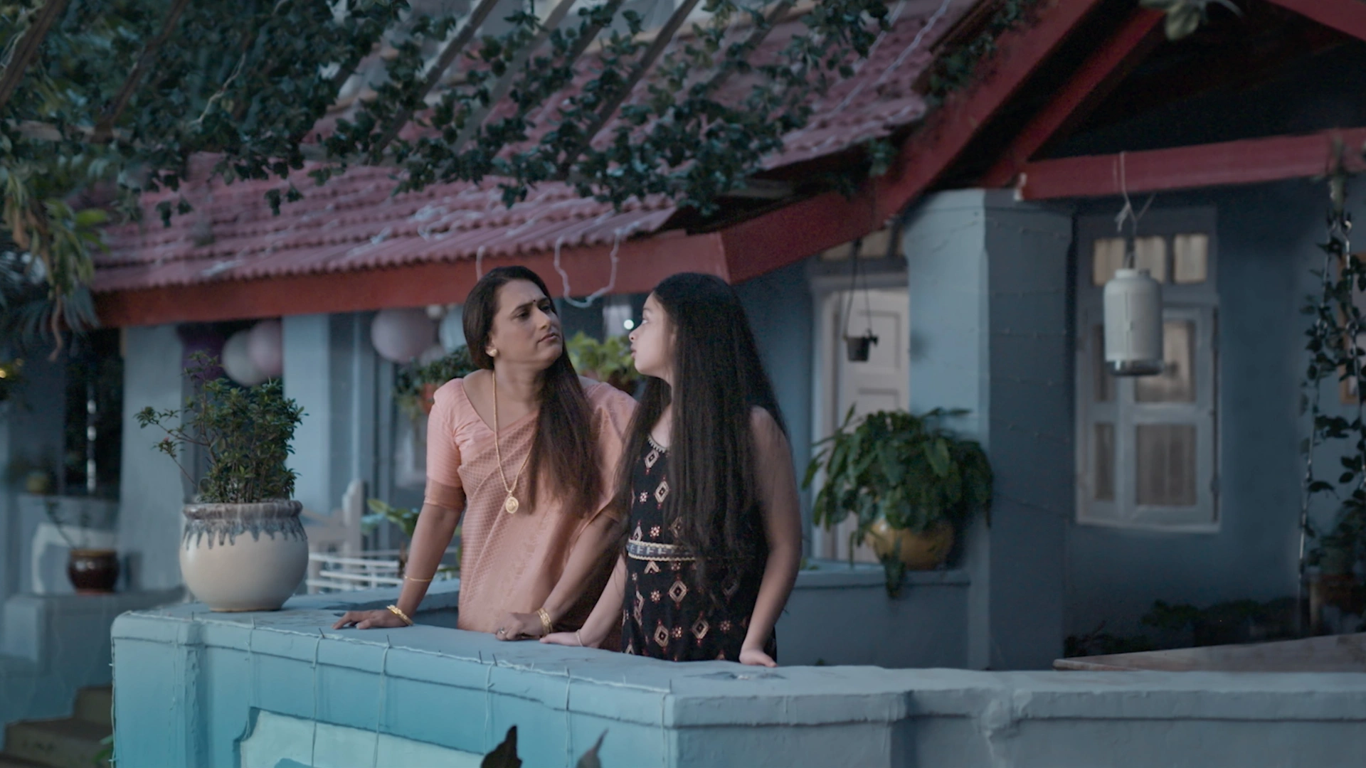 Mother and daughter standing on a blue verandah outside a traditional Goan-style house during evening light in a cinematic family commercial set design.
