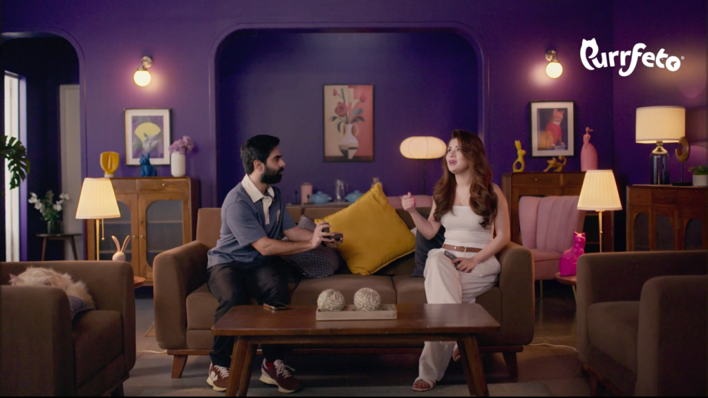 Purrfeto film still showing two characters seated in the purple living-room set with warm practical lamps and timber furniture