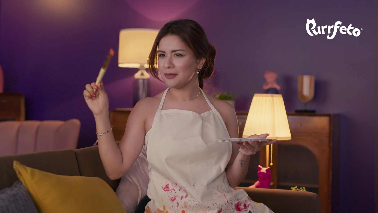 Purrfeto film still showing an artist character seated in the purple living-room set with a mustard cushion and warm practical lamps